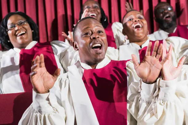 A group of mature African American women and men wearing robes, singing in a church choir. The focus is on the man in the foreground, hands raised, performing with a joyful expression.