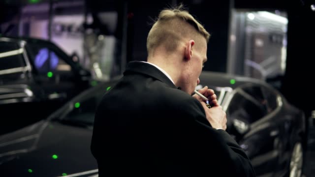 Young man in a black suit with a bow-tie smoking a cigarette by the black car at night.