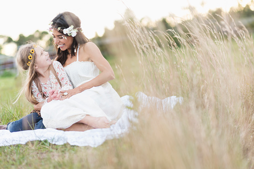A boho-chic young woman and little girl sitting together outdoors in a field of grass, looking at each other, smiling.