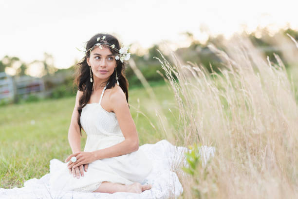 A beautiful, young boho-chic Hispanic woman sitting in a grassy field, wearing a white dress.