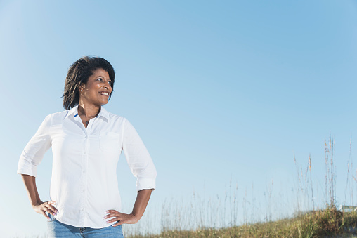 Portrait of a mature African American woman standing outdoors, hands on her hips, smiling with confidence. Clear blue sky in the background.