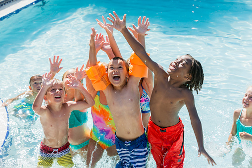A multi-ethnic group of children, 5 to 7 years old, playing in a swimming pool. They are raising their arms, looking up, getting ready to catch something.