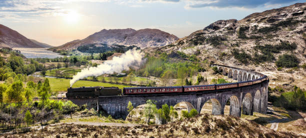 グレンフィナン湖に沈む夕日に対してジャコバイト蒸気機関車列車とスコットランドの鉄道高架橋 - 高架橋 ストックフォトと画像