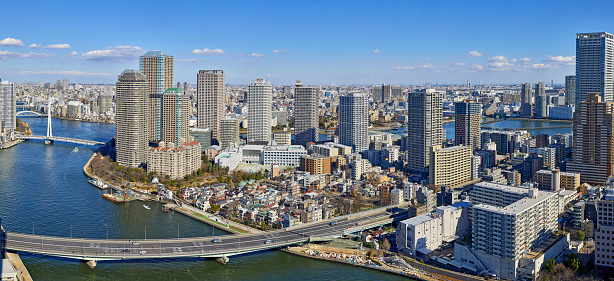 A wide angle panoramic view of Sumida river under blue winter sky in Tokyo with wavy water, boats, bridges and skyscrapers from above.