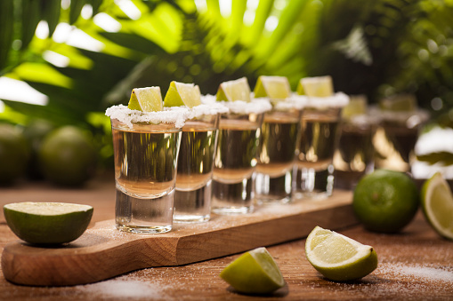 Gold tequila shots with lime fruits on wooden and green tropical leaves background