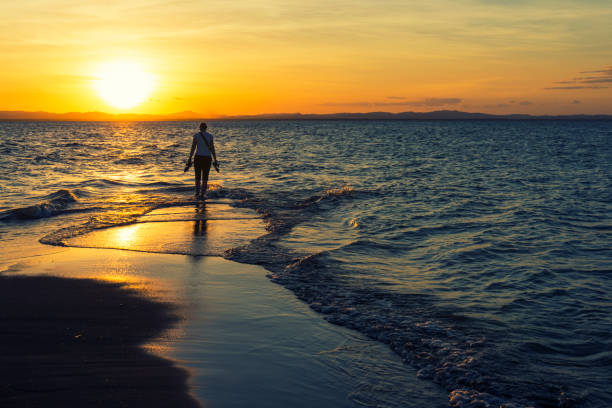 View on narrow part of black diminishing sandy beach  at sunset. Vawes washing the sand from two sides of the coast. No people and no plants, just clear romantic sky. Young woman standing at the end of a sandy beach and contemplating sunset
