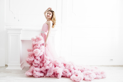 Gorgeous woman portrait in a long pale pink ruffled dress train in a white classic style room. Elegant femininity
