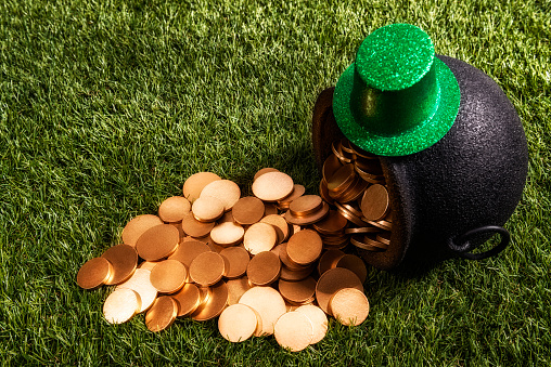 Looking down on a black wrought iron pot of gold  coins that has been knocked over in the grass, with a little green glittery Leprechaun's top hat sitting on top.