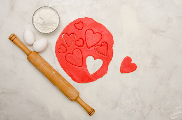 Red dough with a rolling pin and cut out hearts on a white table, top view