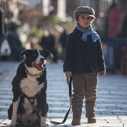 Portrait of little boy with his Border Collie dog on the street.Boy is holding peat leash and he is wearing a black coat,blue scarf,brown baker boy cap,yellow boots and brown trousers.They are seen in full length.Background is blurred and people on the street are seen blurred.Light source is sun and back lit is used.Shot in outdoor with a full frame DSLR camera and a tele lens.