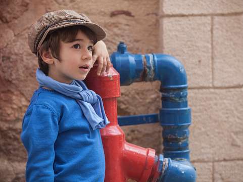 Head and shoulders portrait of little boy wearing a blue sweater,scarf and a brown flat cap.He is standing by a red and blue color fire hydrant.Shot in day light with a full frame DSLR camera.Horizontal framing.