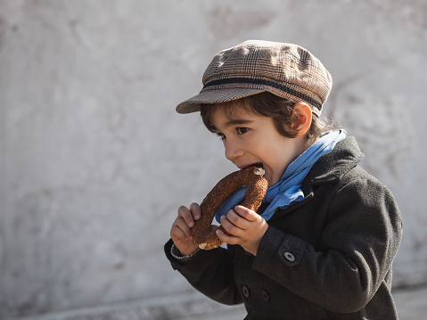 Portrait of little boy wearing a black overcoat,blue scarf and brown baker boy cap eating sesame bagel.He is sitting in front of a beige wall.Background is blurred.Shot in outdoor day light with a full frame DSLR camera.Horizontal composition.