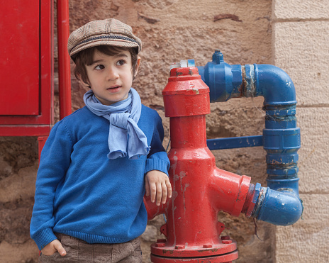Head and shoulders portrait of little boy wearing a blue sweater,scarf and a brown flat cap.He is standing by a red and blue color fire hydrant.Shot in day light with a full frame DSLR camera.Horizontal framing.