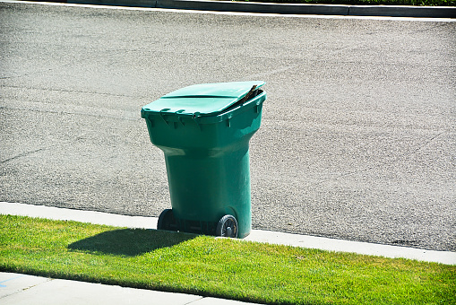 A green household garbage can sits at a curb on a residential street, ready to be picked up by a city garbage truck.