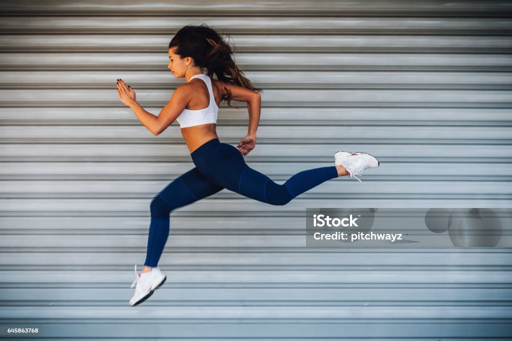 Young female athlete woman running jumping Image of young female athlete running and jumping outdoors. Copy space has been left, shoot with available light. Exercising Stock Photo