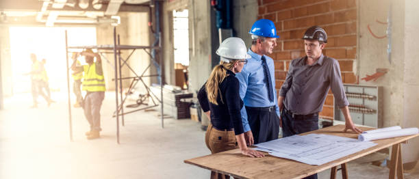 Businessman talking to architects at construction site Three people standing at a table with stretched out plans on it and talking about the construction process and costs. foreman stock pictures, royalty-free photos & images
