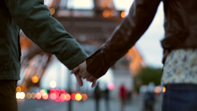 HANDHELD CLOSE UP REAR TRACKING SHOT young man and woman hold hands while walking toward Eiffel Tower in Paris at dusk