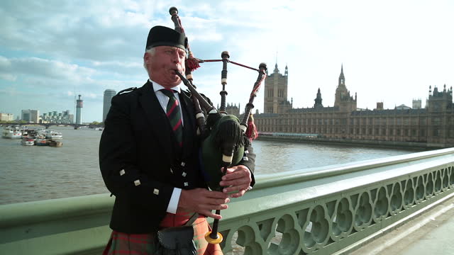 Man in traditional dress plays bagpipes on Westminster Bridge with the Palace of Westminster in the background.