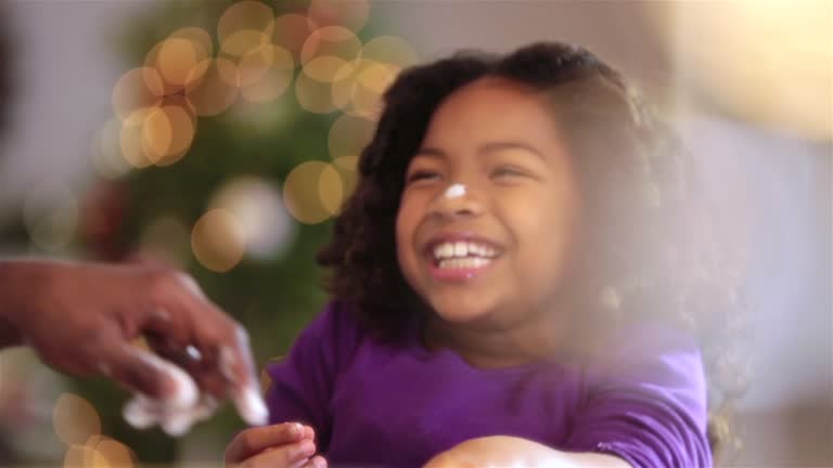 Cute young girl sprinkles flour on cookie dough, father leaves a flour fingerprint on the tip of her nose