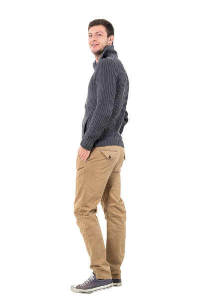 Rear view of young man in jumper smiling at camera Rear view of young man in jumper with hands in pockets smiling at camera. Full body length portrait isolated over white background. turning back stock pictures, royalty-free photos & images