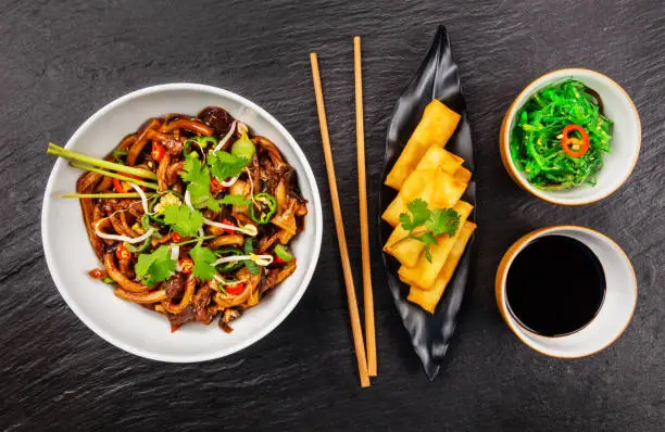 Asian noodles with spicy soy sauce, chicken pieces and spring rolls, top view, served on black stone.