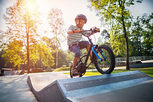 Little boy wearing a helmet is riding a bicycle on ramp in city park on summer day. The boy is aged 6 and is smiling.\n