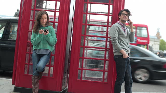 Young man and woman lean on London telephone booths while talking and texting on smartphones.