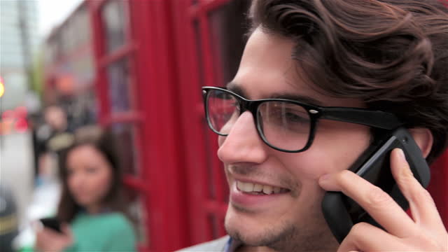 CU. Young man talks on smartphone outside red telephone booth.