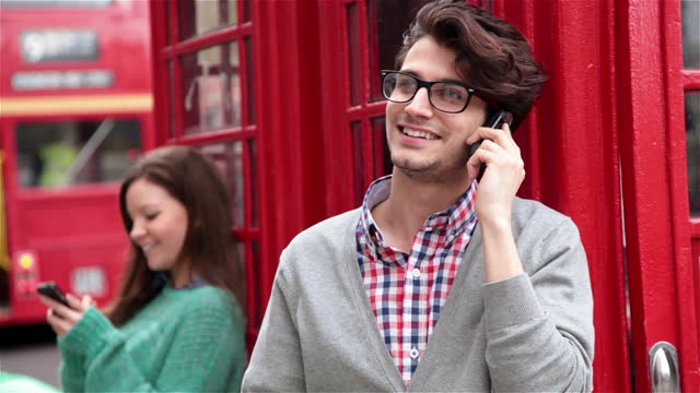 Young man and woman talk and text on smartphones while leaning on red telephone booths.
