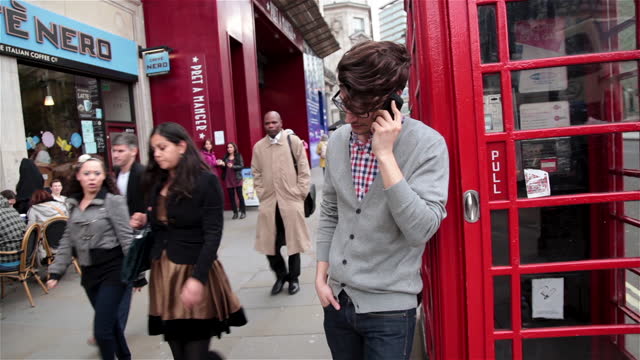 Double decker bus passes as young man leans on telephone booth and talks on smartphone.