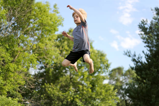 A young boy child is jumping high up by the trees as he play outside on a trampoline on a summer day.