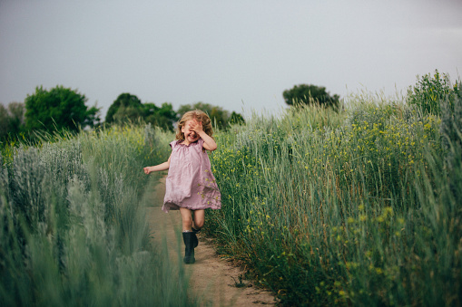 3 year old with long blonde hair, wearing a purple dress in a field of tall high green grass. Holding her hand over her face.