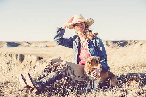 A pretty cowgirl sitting on a grassy prairie, holding her hat with one hand and her little pet dog with the other. She is wearing black cowgirl boots, leather chaps, denim jacket and a white cowgirl hat.