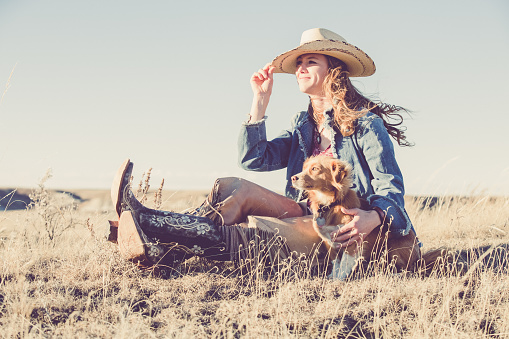 A pretty cowgirl sitting on a grassy prairie, holding her hat with one hand and her little pet dog with the other while looking off into the distance. She is wearing black cowgirl boots, leather chaps, denim jacket and a white cowgirl hat.