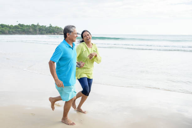 ältere mann und reife frau lachend und joggen am strand - couple senior adult travel action stock-fotos und bilder