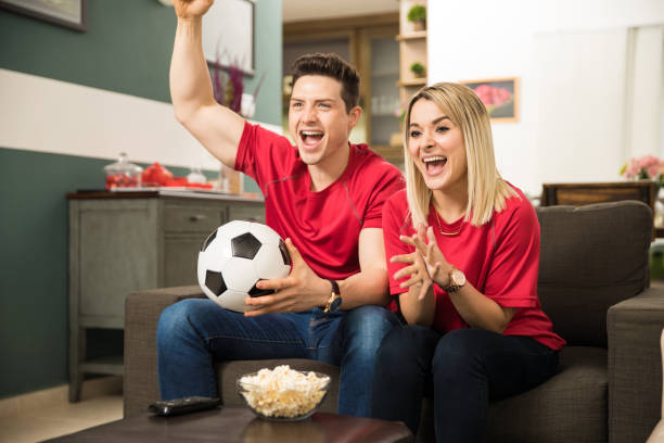 Excited soccer fans watching game Good looking young couple looking surprised and excited while watching a soccer game at home excited football fan cheering for his team on tv stock pictures, royalty-free photos & images