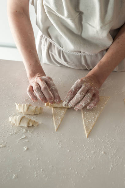 High resolution digital capture of a woman's hands rolling croissant dough on a floured surfce. Woman is wearing a classic baker's apron, and her hands are coated with flour. Two croissants are completed, one is being rolled, and the other is an open and flat triangle of dough.