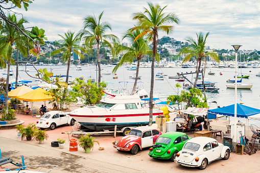 People sit and relax at the waterfront of Acapulco, Mexico with docked boats in the background and parked Volkswagen Beetle cars in the foreground.