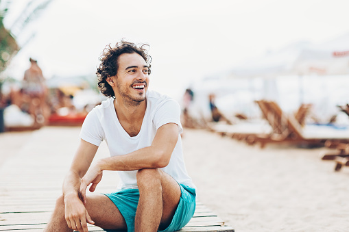 Cheerful young man sitting on the beach in the summer and looking away, with copy space