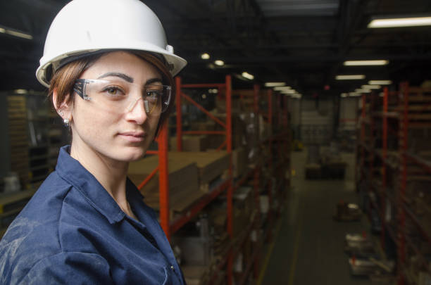 Woman manager standing on mezzanine overlooking her manufacture's warehouse. She wears a white hard hat and a blue sarrau