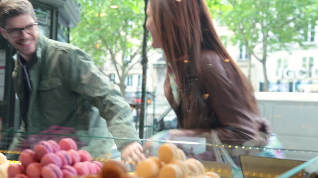 Excited young couple stop to look at colorful macaroons in the window of a French bakery and point out the ones they want.