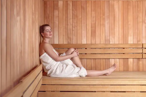 Side view full length portrait of young woman smiling sitting on wooden sofa bench in sauna wrapped in white towel, with hair in donut bun