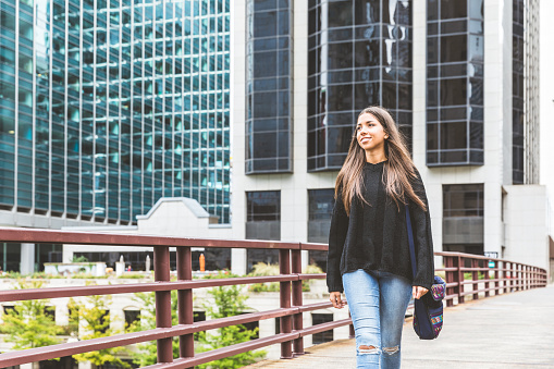 Smiling girl walking in Chicago. Portrait of a young woman, mixed race, looking away from camera, walking over a bridge with modern buildings on background