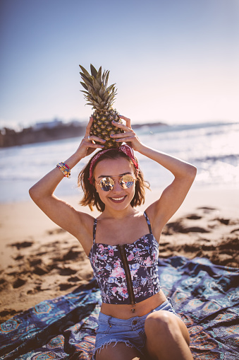 Beautiful young woman in boho style smiling and holding a pineapple at the beach