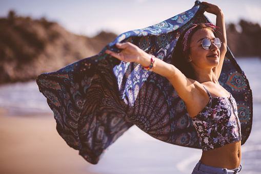 Attractive young woman in boho style, wearing sunglasses holding a multi-colored scarf at the beach