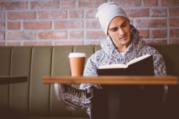 Young smart fashionable man, sitting in coffee shop, drinking coffee, reading a book and smiling in front of brick wall