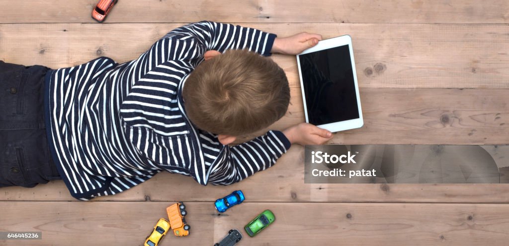 kid on floor with tablet pc Top view header image with child laying down on the floor watching a movie on his tablet instead of playing with his toy cars Child Stock Photo