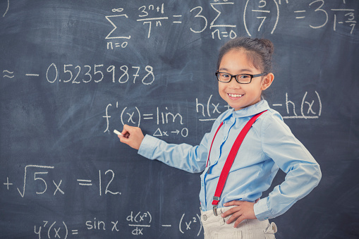 Smart Asian elementary school student smiles while completing math problem on chalkboard in classroom.