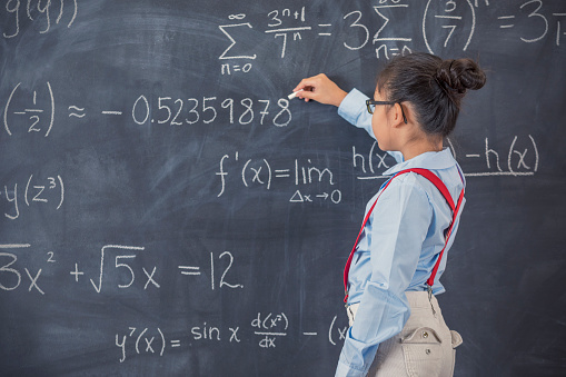 Confident Asian elementary schoolgirl works math problem on chalkboard in classroom.