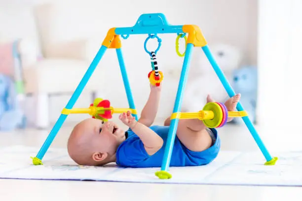 Cute baby boy on colorful playmat and gym, playing with hanging rattle toys. Kids activity and play center for early infant development. Newborn child kicking and grabbing toy in white sunny nursery